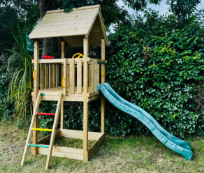 Maisonnette de jeu en bois avec toboggan vert et échelle d'escalade placée à côté d'une haie